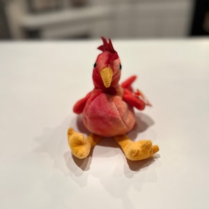 May include: A red and orange plush rooster toy with yellow feet. The toy is sitting on a white surface.