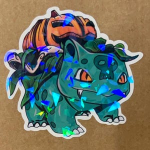 Könnte beinhalten: Ein grüner und blauer holografischer Aufkleber eines Pokemon-Charakters, Bulbasaur, mit einem Kürbis-Jack-o-Lantern auf dem Rücken.