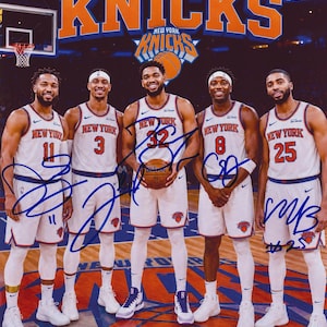 May include: A 2026 New York Knicks limited edition autographed photograph featuring five basketball players in white home uniforms with blue and orange trim, standing on a court, each jersey displaying a unique player number and team name.