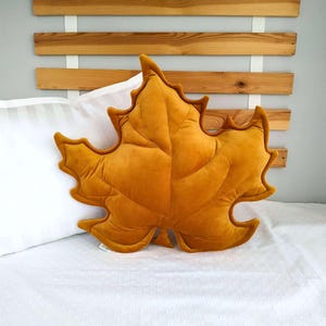 May include: Mustard yellow leaf-shaped pillow. The pillow is made of a soft, velvety material and has a detailed leaf vein pattern. The pillow is resting on a white bed with a striped pillow and a wooden headboard in the background.