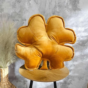May include: A plush, mustard yellow pillow shaped like a four-leaf clover. The pillow is sitting on a wooden table.