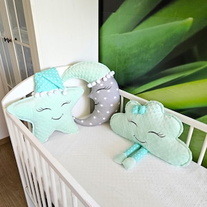 Può includere: Una culla bianca con tre cuscini di peluche: una stella verde menta con un volto addormentato, una luna crescente grigia con stelle bianche e una nuvola verde menta con gambe. I cuscini hanno dettagli di pompon e pois bianchi.