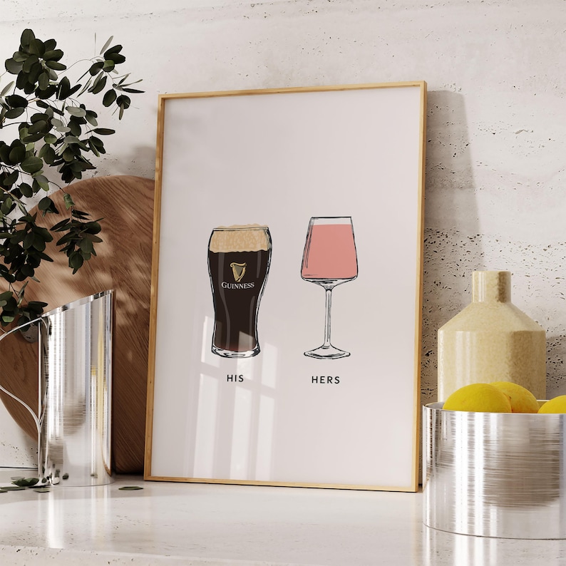 May include: A framed print featuring two illustrated glasses: a pint of Guinness and a stemmed glass with a pink drink. The words "HIS" and "HERS" are printed below the glasses. The print has a gold frame and is displayed on a white surface.