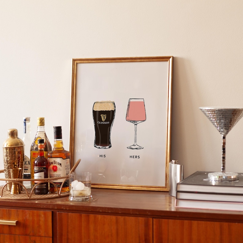 May include: A framed print with a gold border featuring illustrations of a pint of Guinness and a pink cocktail glass, labeled "HIS" and "HERS". The print is displayed on a wooden cabinet with bar accessories.