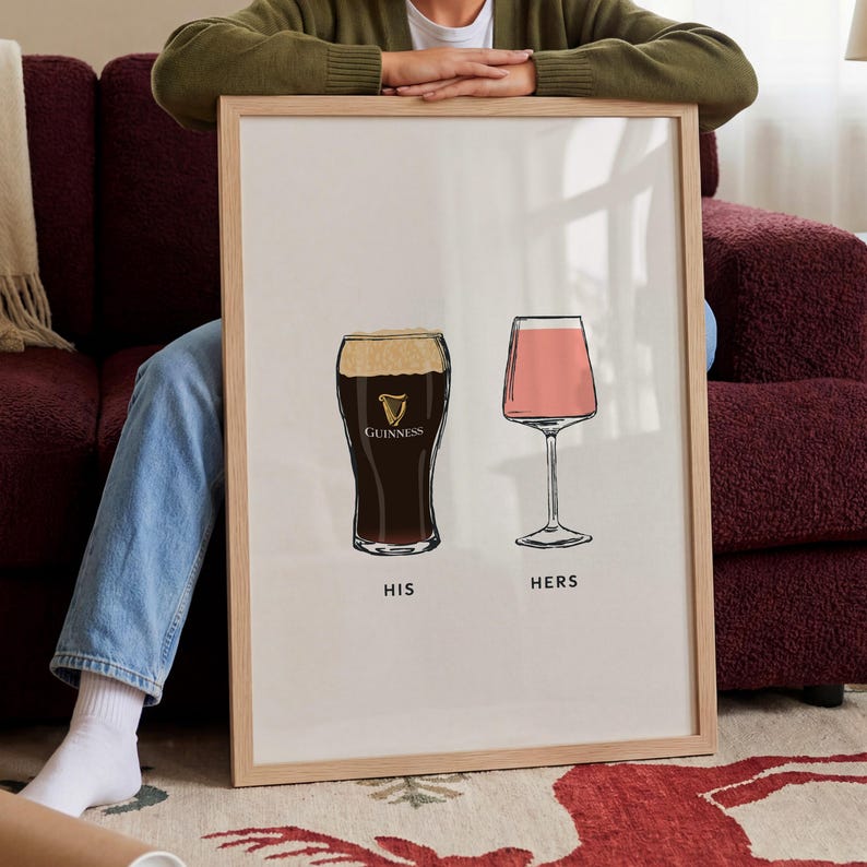May include: Framed art print featuring two illustrated glasses: a pint of Guinness and a pink cocktail. The words "HIS" and "HERS" are printed below the glasses. The print is in a light wood frame.