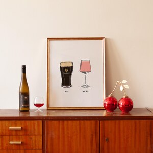 May include: A framed print with illustrations of a pint of Guinness and a pink cocktail glass, labeled "HIS" and "HERS". A wine bottle and a small glass of red wine are on the left. Two red decorative spheres with gold leaves are on the right.