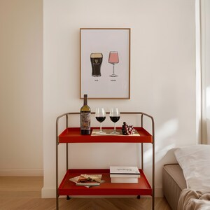 May include: A red serving cart with two shelves holds a wine bottle, two wine glasses, and a small wooden tray with grapes. A framed print above displays two stylized glasses with the words "HIS" and "HERS".