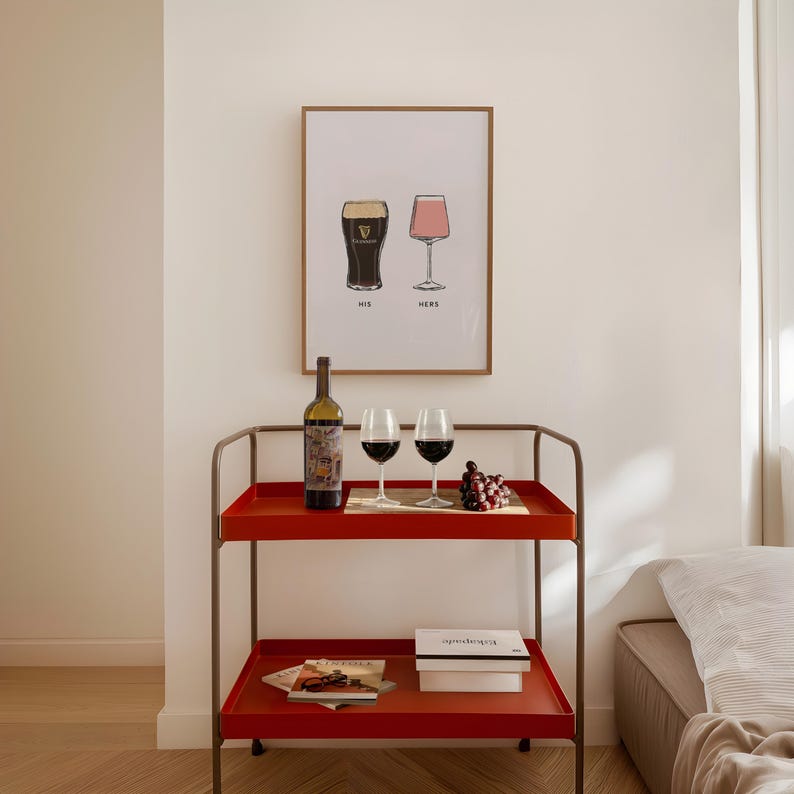 May include: A red serving cart with two shelves holds a wine bottle, two wine glasses, and a small wooden tray with grapes. A framed print above displays two stylized glasses with the words "HIS" and "HERS".