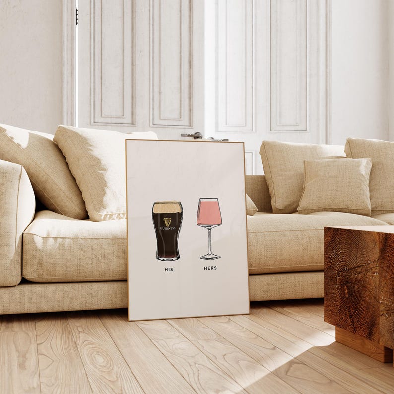 May include: Framed art print featuring a pint of Guinness and a pink wine glass, labeled "HIS" and "HERS" respectively. The print is displayed on a wooden floor, leaning against a beige sofa. The artwork has a minimalist design.