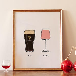 May include: A framed print with a gold border features illustrations of a pint of Guinness and a wine glass with pink liquid. The words "HIS" and "HERS" are below the glasses. A bottle of wine, a small glass of red wine, and two red disco ball ornaments are in the foreground.