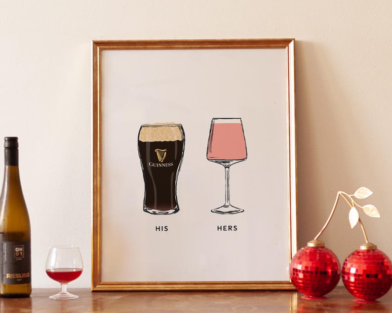 May include: A framed print with a gold border features illustrations of a pint of Guinness and a wine glass with pink liquid. The words "HIS" and "HERS" are below the glasses. A bottle of wine, a small glass of red wine, and two red disco ball ornaments are in the foreground.