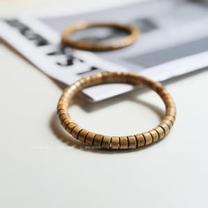 Może przedstawiać: Dwie drewniane bransoletki z koralików. Bransoletki wykonane są z małych, prostokątnych drewnianych koralików nawleczonych razem. Bransoletki są w kolorze jasnobrązowym i spoczywają na białej powierzchni. Średnica ok. 7 cm.