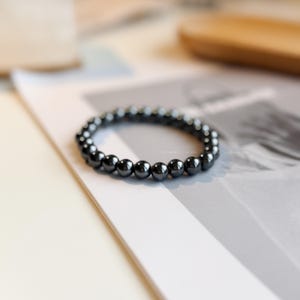 May include: A dark grey beaded bracelet. The bracelet is made of small, round, polished beads strung together. The beads have a metallic sheen and reflect light. The bracelet is resting on a white and grey surface.