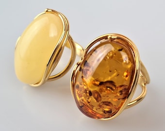May include: Two oval-shaped amber rings with gold settings. The ring on the left has a light yellow amber stone, while the ring on the right has a darker amber stone.