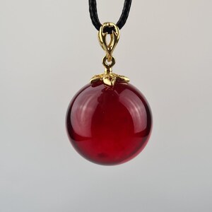 May include: A red glass ball pendant on a black cord. The pendant has a gold-colored metal top.