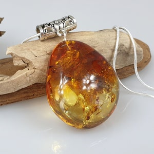 Puede incluir: Collar con colgante de ámbar en forma de lágrima y cadena de plata. El ámbar muestra tonos dorados y translúcidos, con inclusiones visibles. La fianza plateada tiene un diseño floral. El collar descansa sobre madera flotante.