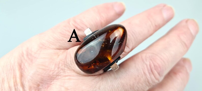 K&ouml;nnte beinhalten: Ein Ring mit einem gro&szlig;en, ovalen Bernstein, der in eine silberne Fassung gefasst ist. Der Stein ist dunkelbraun mit goldenen Einschl&uuml;ssen.