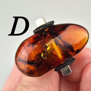 K&ouml;nnte beinhalten: Ein Ring mit einem gro&szlig;en, ovalen Bernstein, der in eine silberne Fassung gefasst ist. Der Bernstein ist tief orangefarben mit schwarzen und wei&szlig;en Einschl&uuml;ssen. Die Fassung ist mit zwei schwarzen B&auml;ndern verziert.