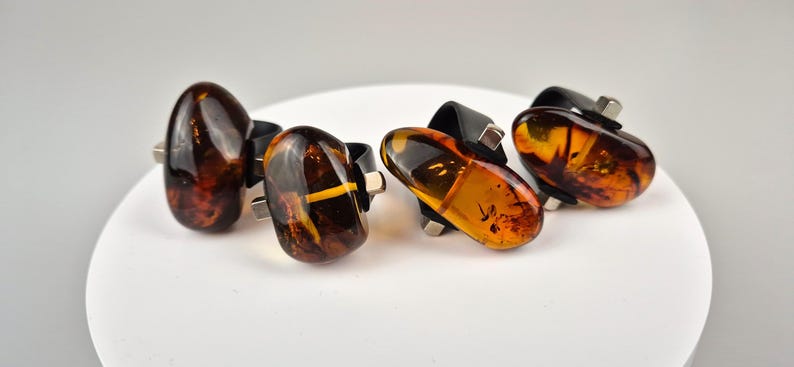 K&ouml;nnte beinhalten: Vier Bernsteinringe mit schwarzen Fassungen. Jeder Ring zeigt ein anderes St&uuml;ck Bernstein mit Einschl&uuml;ssen. Der Bernstein hat eine warme braune Farbe mit Gelb- und Oranget&ouml;nen.