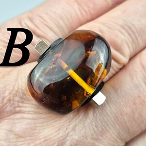 K&ouml;nnte beinhalten: Nahaufnahme eines braunen Glasrings mit einem gelben Streifen. Der Ring befindet sich an einem Finger. Der Buchstabe "B" ist im Hintergrund sichtbar.