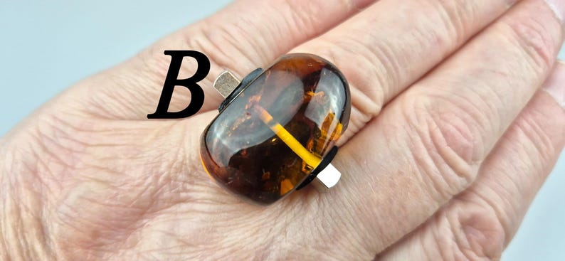 K&ouml;nnte beinhalten: Nahaufnahme eines braunen Glasrings mit einem gelben Streifen. Der Ring befindet sich an einem Finger. Der Buchstabe "B" ist im Hintergrund sichtbar.