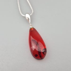 May include: A teardrop-shaped pendant made of translucent deep red amber with dark internal inclusions, suspended from a polished silver-toned metal bail and a matching silver-toned snake chain necklace, showcasing a smooth, polished surface finish.