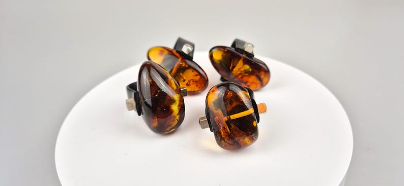 K&ouml;nnte beinhalten: Vier Bernsteinringe mit schwarzen Metallfassungen. Die Ringe liegen auf einer wei&szlig;en Oberfl&auml;che.