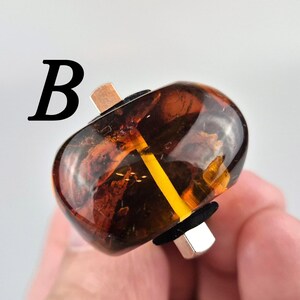 K&ouml;nnte beinhalten: Nahaufnahme einer braunen und bernsteinfarbenen Glasperle mit einem goldenen Metallband und einem schwarzen Gummiband. Die Perle wird von einer Hand gehalten.