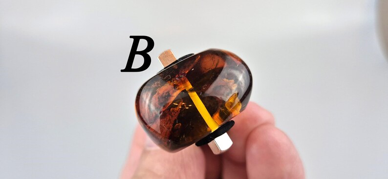 K&ouml;nnte beinhalten: Nahaufnahme einer braunen und bernsteinfarbenen Glasperle mit einem goldenen Metallband und einem schwarzen Gummiband. Die Perle wird von einer Hand gehalten.