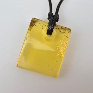 Peut inclure: Un pendentif rectangulaire en ambre sur un cordon noir. Le pendentif est jaune translucide avec des éclats d'or et de brun foncé. Le cordon est noué en haut, et le pendentif a un trou pour le passage du cordon.