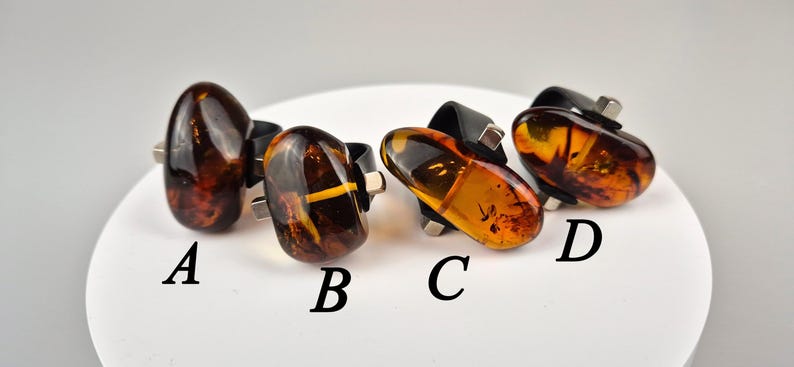K&ouml;nnte beinhalten: Vier Bernsteinringe mit unterschiedlichen Einschl&uuml;ssen, gefasst in Silber und schwarzem Metall. Die Ringe sind mit A, B, C und D gekennzeichnet.