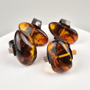 May include: Four amber rings with black metal settings. The rings are arranged on a white surface.
