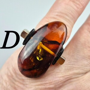 K&ouml;nnte beinhalten: Ein Ring mit einem gro&szlig;en, ovalen Bernstein, der in eine silberne Fassung gefasst ist. Der Stein ist tief orangefarben mit goldenen und braunen Streifen. Die Fassung ist schlicht und modern, mit einem schwarzen Lederakzent auf jeder Seite des Steins.