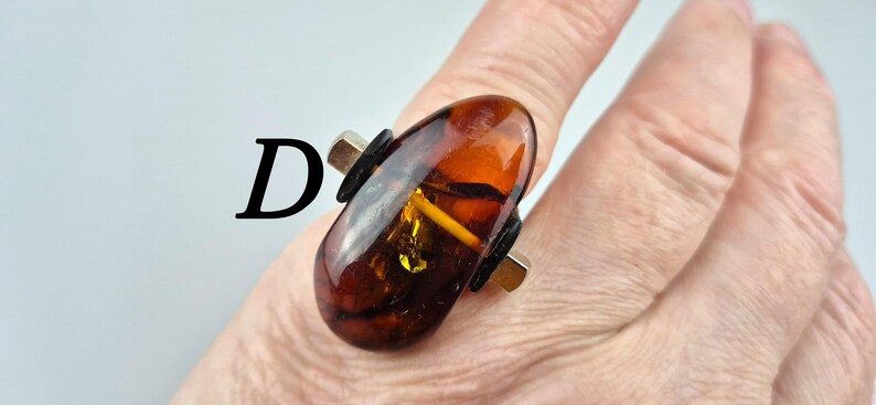 K&ouml;nnte beinhalten: Ein Ring mit einem gro&szlig;en, ovalen Bernstein, der in eine silberne Fassung gefasst ist. Der Stein ist tief orangefarben mit goldenen und braunen Streifen. Die Fassung ist schlicht und modern, mit einem schwarzen Lederakzent auf jeder Seite des Steins.