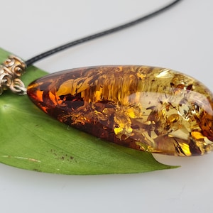 Può includere: Un ciondolo in ambra a forma di lacrima con un cordoncino in pelle nera. L'ambra è una miscela di marrone e giallo con inclusioni. Il ciondolo è attaccato al cordoncino con una chiusura in metallo argentato.