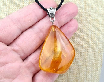 May include: A teardrop-shaped amber pendant with a silver bail, hanging from a black cord. The pendant is a deep orange color and has a smooth, polished finish.
