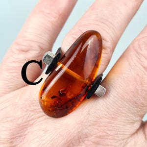 K&ouml;nnte beinhalten: Ein silberner Ring mit einem gro&szlig;en, bernsteinfarbenen Stein, der in einem einzigartigen, asymmetrischen Design gefasst ist. Der Stein ist in einem tiefen, satten Braun mit orangefarbenen und gelben Nuancen.