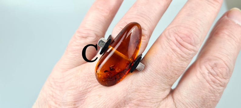K&ouml;nnte beinhalten: Ein silberner Ring mit einem gro&szlig;en, bernsteinfarbenen Stein, der in einem einzigartigen, asymmetrischen Design gefasst ist. Der Stein ist in einem tiefen, satten Braun mit orangefarbenen und gelben Nuancen.