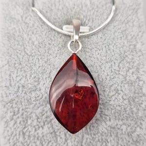 Puede incluir: Collar de plata con un colgante en forma de lágrima de piedra roja translúcida. El colgante está suspendido de una cadena de plata. La piedra tiene una superficie pulida. El collar se muestra sobre una superficie gris texturizada.