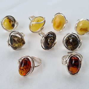 Op de afbeelding: Een verzameling van acht amber ringen met zilveren draadbevestigingen. De ringen hebben verschillende tinten amber, waaronder geel, bruin en groen.