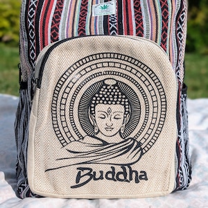 Op de afbeelding: Een kleurrijke rugzak met een Boeddha-ontwerp. De tas heeft een beige voorvak met een zwarte Boeddha-illustratie en het woord "Boeddha". De boven- en zijkanten zijn van stof met rode, zwarte en witte patronen. Een klein groen label is zichtbaar.
