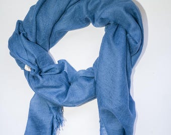 May include: A blue scarf with a fringed edge. The scarf is made of a soft material and is draped in a loop. The scarf is a solid color and appears to be lightweight.