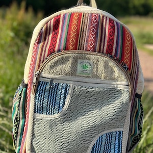 Puede incluir: Una mochila colorida con un patrón de rayas rojas, azules y blancas. La bolsa tiene un bolsillo frontal con un diseño de cheurón azul y gris y un bolsillo más pequeño con rayas azules. El texto "Himalayan Hemp THC Free" es visible.