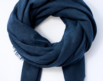 May include: A navy blue scarf with a soft, textured appearance. The scarf is draped in a circular shape, with the ends hanging down. The edges of the scarf have a fringed detail. The scarf is made of a soft material.