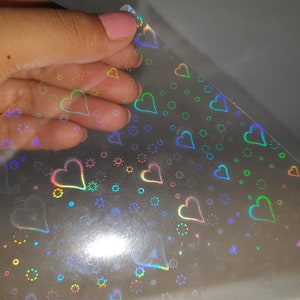 May include: A sheet of iridescent holographic paper with a repeating pattern of hearts and circles. The paper is translucent and has a rainbow sheen.