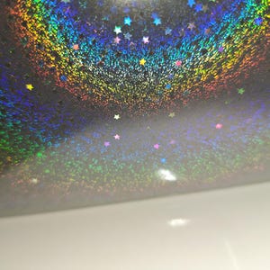 May include: A sheet of holographic material featuring a dark background covered in a dense pattern of small, multi-coloured stars. The surface reflects light in a spectrum of rainbow colours, creating a shimmering effect across the entire surface.