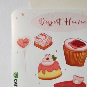 May include: A sticker sheet with a pink and white color scheme featuring various desserts, including a heart-shaped lollipop, a cupcake with pink frosting and a cherry, a square cake with pink and white swirls, a cupcake with pink frosting and raspberries, a popsicle with pink and white stripes, and two pink and white candy squares. The sticker sheet has the text "Dessert Heaven" in pink lettering.