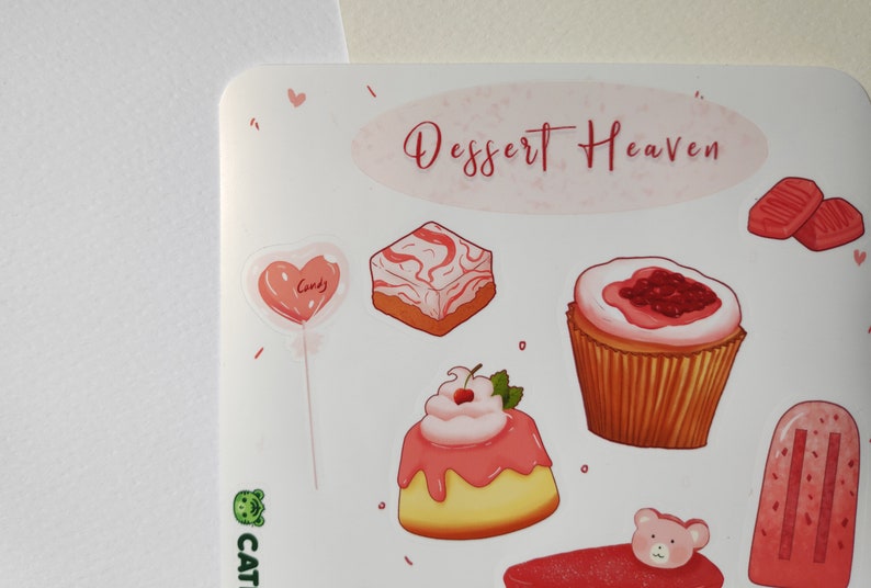 May include: A sticker sheet with a pink and white color scheme featuring various desserts, including a heart-shaped lollipop, a cupcake with pink frosting and a cherry, a square cake with pink and white swirls, a cupcake with pink frosting and raspberries, a popsicle with pink and white stripes, and two pink and white candy squares. The sticker sheet has the text "Dessert Heaven" in pink lettering.