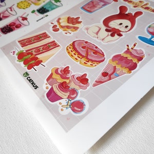 Puede incluir: Una hoja de pegatinas con varios postres de dibujos animados lindos y un conejito con un gorro rojo. Las pegatinas est&aacute;n sobre un fondo rosa y blanco con un patr&oacute;n de cuadr&iacute;cula. El texto "Catkus" est&aacute; impreso en la esquina inferior izquierda.
