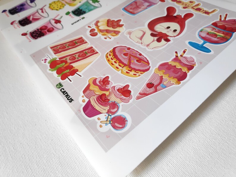 Puede incluir: Una hoja de pegatinas con varios postres de dibujos animados lindos y un conejito con un gorro rojo. Las pegatinas est&aacute;n sobre un fondo rosa y blanco con un patr&oacute;n de cuadr&iacute;cula. El texto "Catkus" est&aacute; impreso en la esquina inferior izquierda.